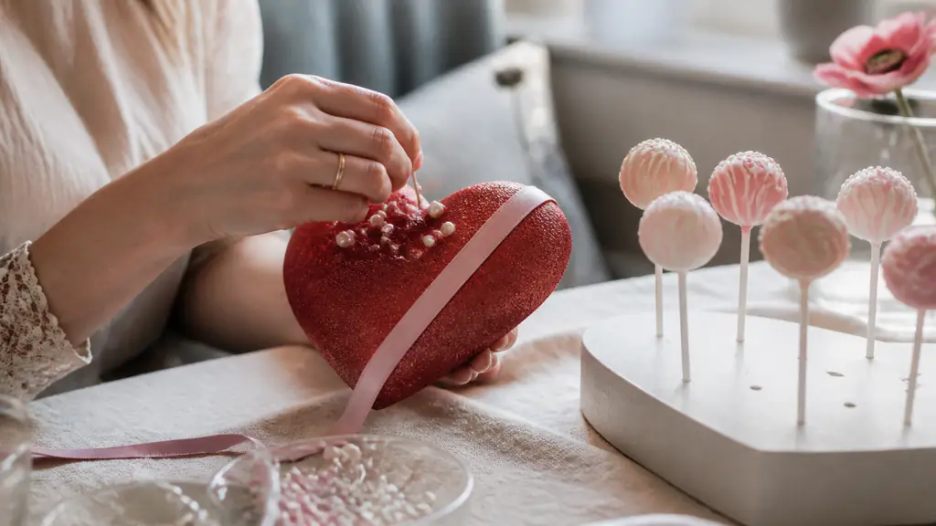 Editorial close-up of hands decorating a red polystyrene heart with pearl-headed sequins and a pink ribbon, beside a heart-shaped cake pop stand on a romantic table, soft natural light, no text, no logo